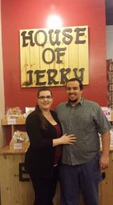 A male and a femal in front of a wooden House of Jerky sign in a store